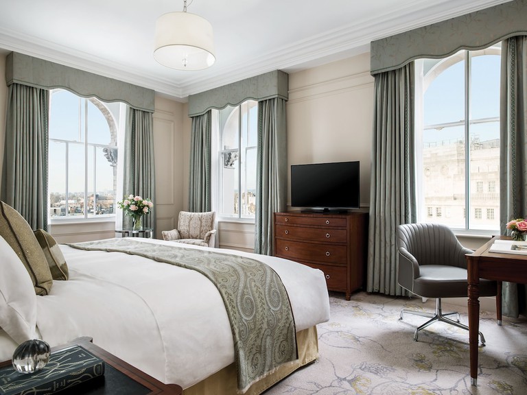 A luxury king bedroom in light beige and gold tones with wall of windows at the Langham in London