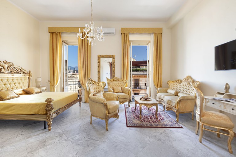 Plush gilded furniture, including a bed, armchairs, couch, and desk with chair, in a hotel room at Relais Antica Napoli; there is a view of buildings from the balcony
