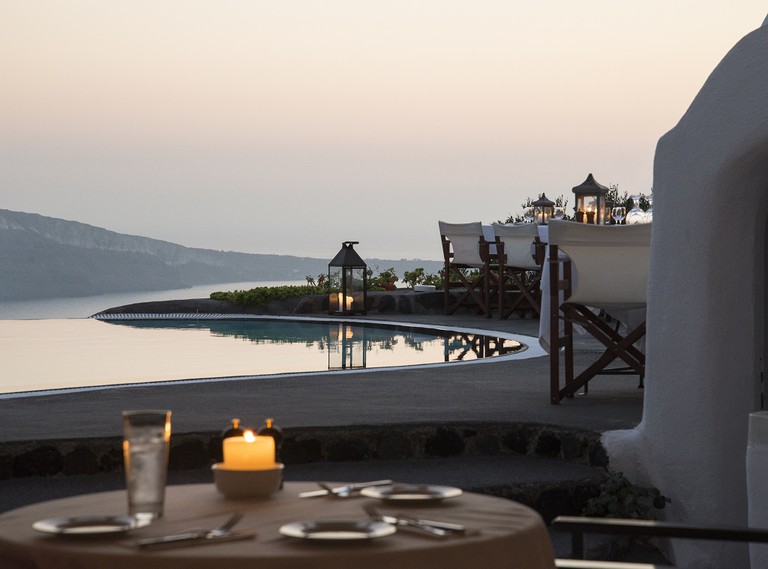A poolside dining terrace with candles lit on tables and by the side of the pool overlooking the sea at sunset in Santorini.