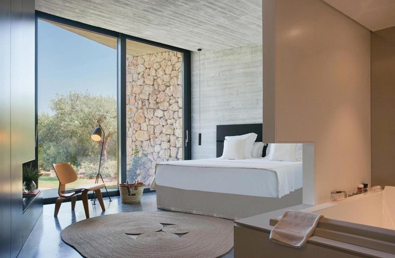 A light ensuite bedroom at Son Brull Hotel and Spa, with large windows, a lamp, plant and a wooden chair
