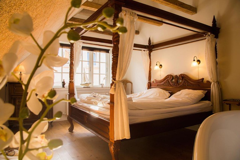 Cosy bedroom with carved-wood four-poster bed, exposed-beam ceiling, quaint lamps and white orchids at Postgården Holsted
