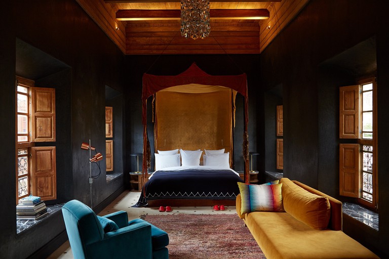 A dark-walled guest room at El Fenn, Marrakech with a four-poster bed, traditional wooden shutters and colourful suede armchairs.