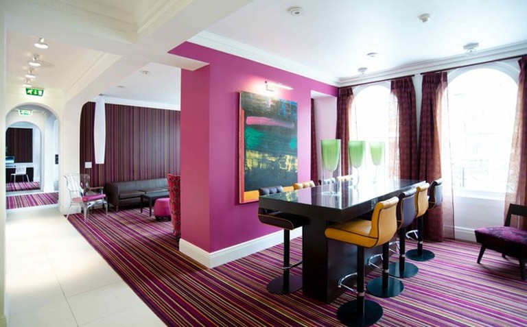 Lounge with magenta partition wall, high-top table and striped magenta carpeting at Safestay Elephant and Castle, London