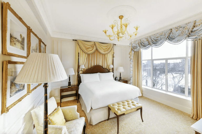 An elegant room at the Savoy with a gold chandelier, draped curtains, a curtained headboard, gilt-framed artwork and a view of the River Thames through a large window.