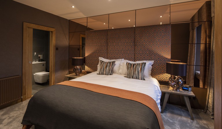 A chic en-suite bedroom in mauve hues with a queen bed, patterned headboard, mirrored wall and plush carpet at the Heathmount