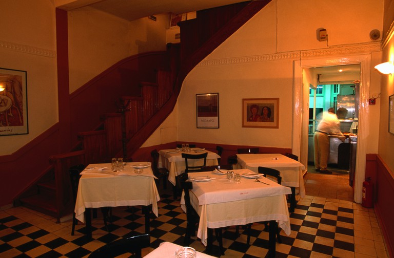 Un comedor interior en el restaurante Vlassis con suelo de baldosas a cuadros, mesas con manteles blancos y una escalera de madera