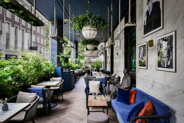 Outdoor terrace at the Churchill Bar & Terrace, with hanging plants, blue and grey seating, a marble wall with photographs of Churchill, and a seated metal statue of the man himself.