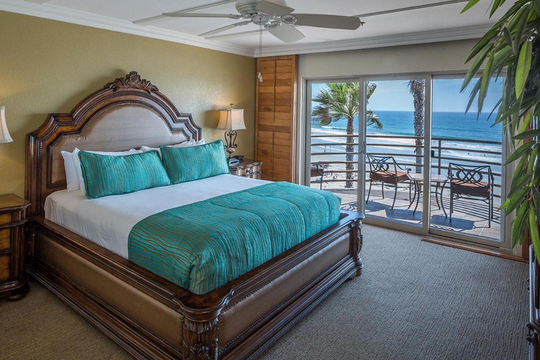 Elegant ocean-view bedroom at Pacific Terrace Hotel featuring a grand wooden-frame bed and a private balcony overlooking the beach