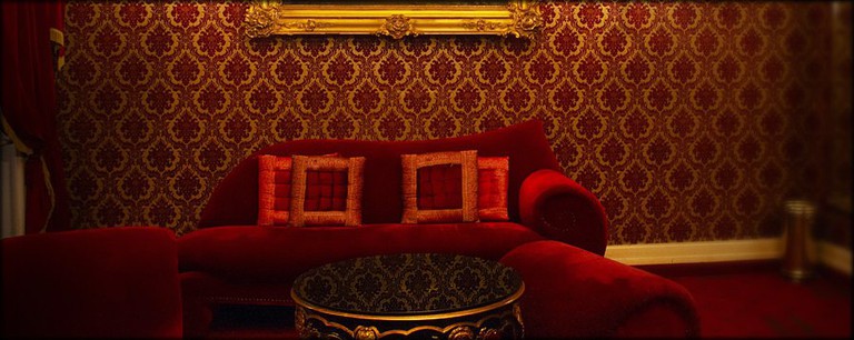 Un salón oscuro con papel tapiz estilo brocado flocado y un sofá de terciopelo rojo intenso.