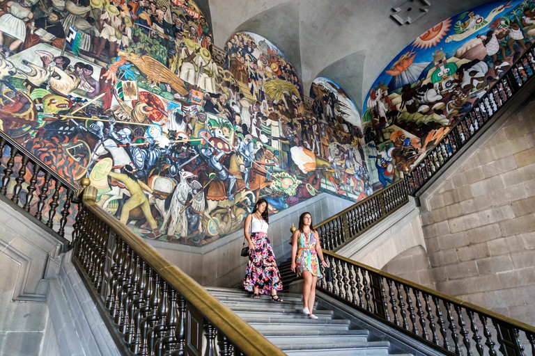 Two tourists walk down the main stairwell in the Palacio Nacional, The History of Mexico mural by Diego Rivera behind them