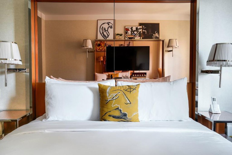 A crisp double bed with a mirrored headboard and a yellow patterned pillow facing a flat screen TV in a room at the Mondrian Park Avenue