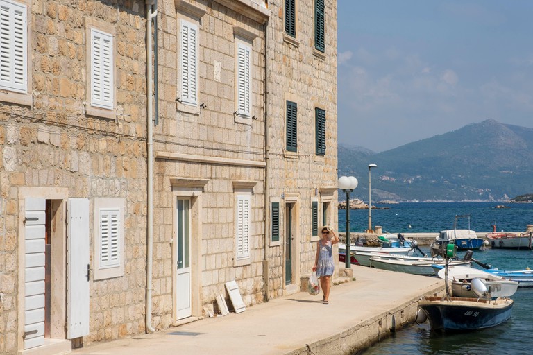 A woman in a blue sundress strolls along the seafront promenade in Šipan, past small boats and traditional stone houses with shuttered windows.