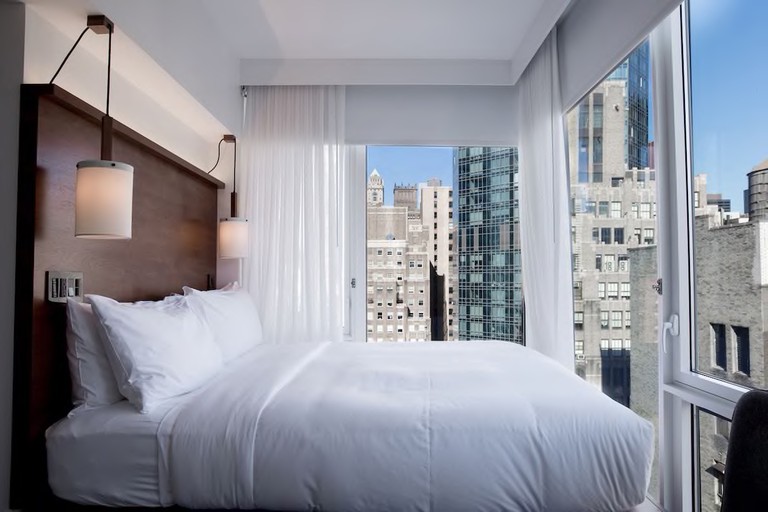 A bright bedroom at Arlo NoMad, with a large bed, bedside pendant lamps and floor-to-ceiling windows on two sides with city views