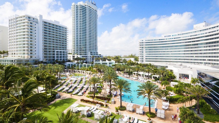 An outdoor pool and palm trees, and the white hotel buildings of Fontainebleau Miami Beach