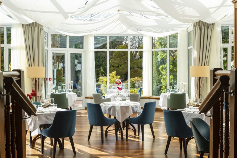 A dining room at Kingsmills Hotel Inverness with small tables and white table cloths, as well as floor-to-ceiling windows with white curtains overlooking green gardens