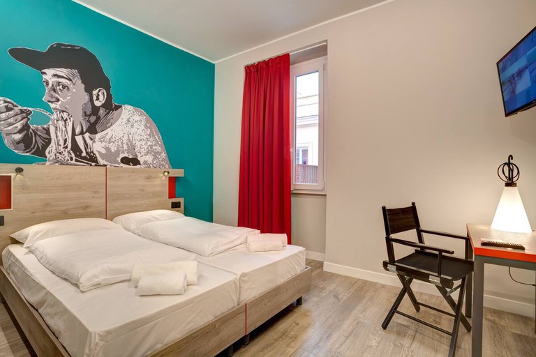 A double bed and a desk in a guest room at MEININGER Roma Termini; there is artwork of a man eating spaghetti on a turquoise wall