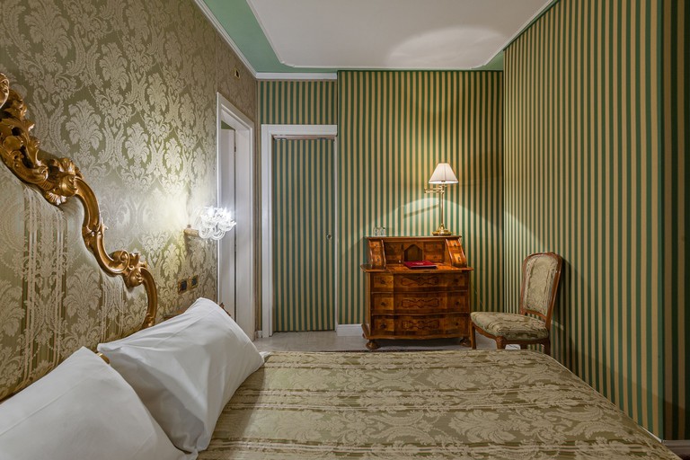 Compact double room at the Hotel Al Ponte Antico, Venice, with walls and furnishings decorated in a mixture of green striped fabric and cream fleurs-de-lys patterns.