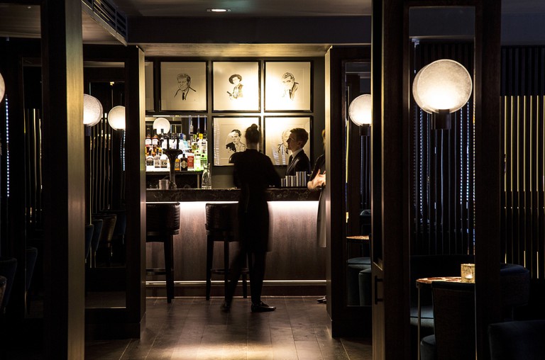 Two people stand at the dimly lit, dark-wood bar in Dakota Glasgow