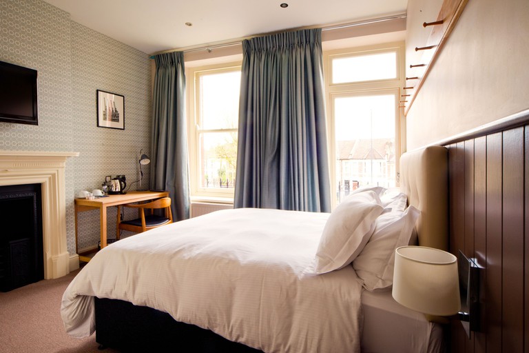 Large double bed with white sheets opposite a TV and desk with kettle and mugs in an airy, comfy room at The Wellington.