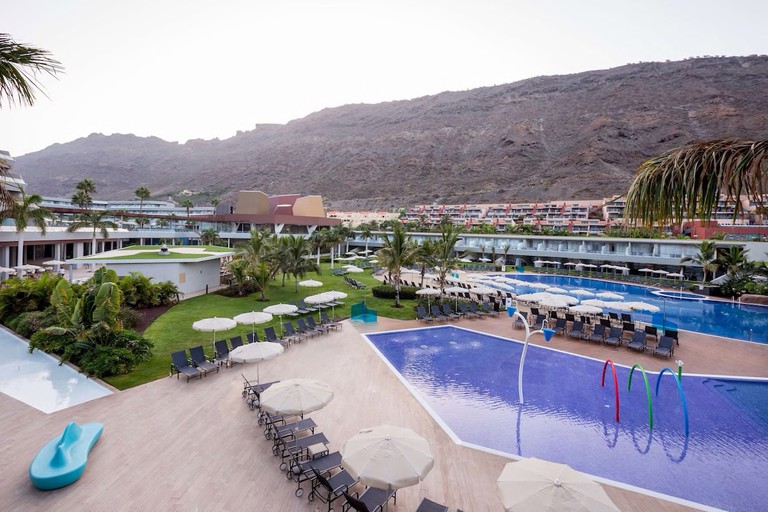 Two outdoor pools at Radisson Blu Resort Gran Canaria surrounded by sun loungers, with buildings and a brown mountain in the background