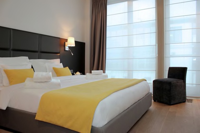 Large double bed with white sheets and yellow cushions in sleek, minimal room with large windows at H15 Boutique.