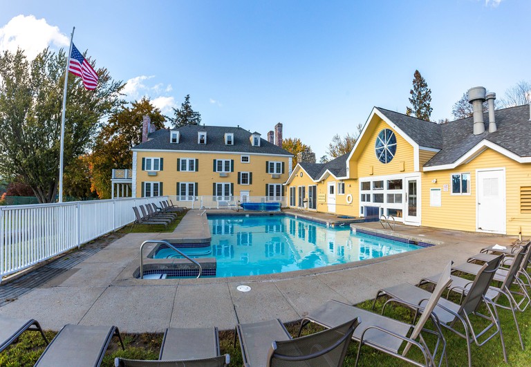 The yellow exterior of Bethel Inn Resort with an outdoor swimming pool and lounge chairs and an American flag flying on a tall flagpole