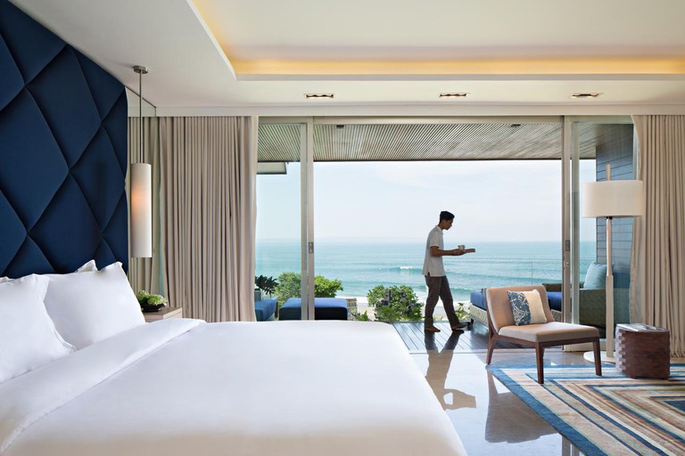 A man walking with a tray bedroom and balcony with a view of the ocean at Como Uma Canggu