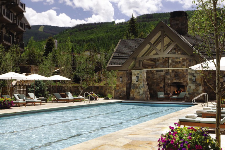 The outdoor pool at Four Seasons Resort Vail with lounge chairs and a fireplace backed by green mountains