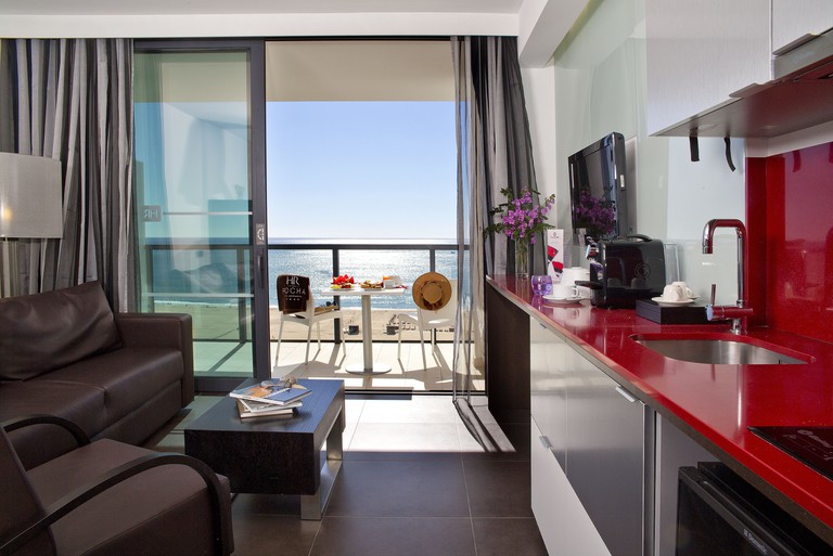 A modern room at Hotel da Rocha with a sofa, armchair, coffee table, TV, red kitchen worktop, sink and balcony with sea views