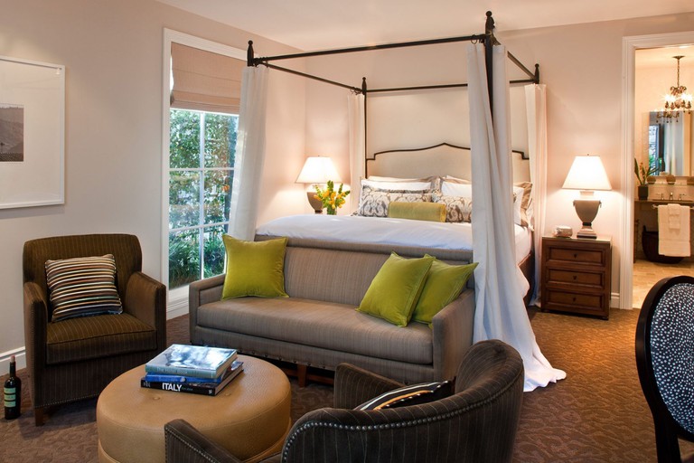 A room at Hotel Yountville with white walls, a four-poster double bed surrounded by white curtains, and a brown sofa seating area scattered with lime cushions.
