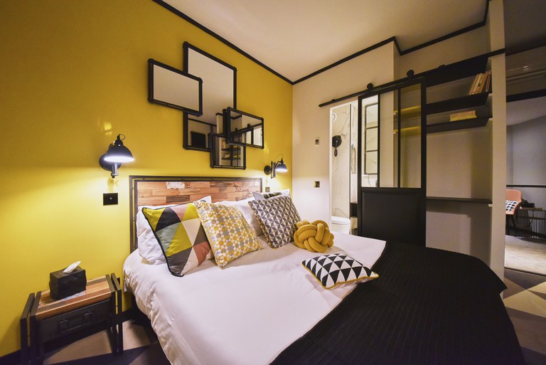 Double bed with patterned cushions in stylish room with a collection of mirrors above the bed, sliding door leading to the bathroom and bookshelves at Hotel Madrigal.