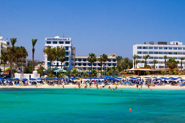 Turquoise sea with blue parasols on beach and Anonymous Beach Hotel in the background