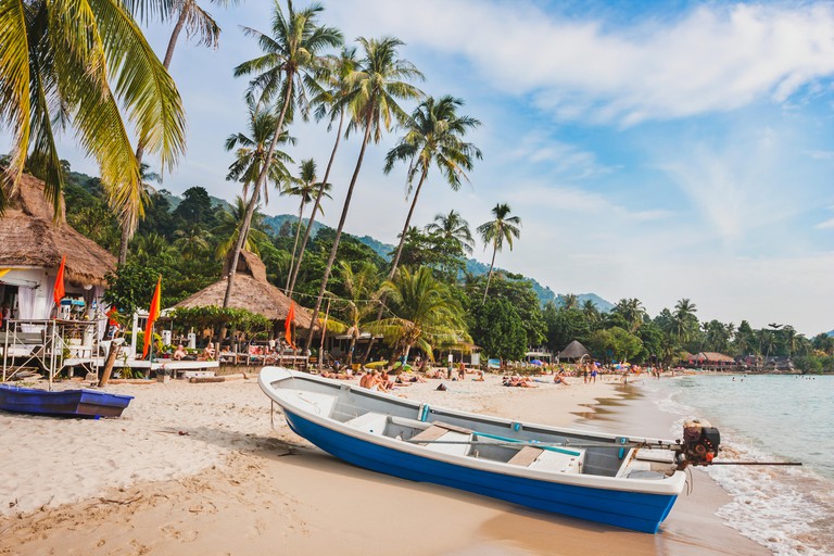 A beautiful tropical beach in Thailand with a wooden boat, thatched buildings and palm trees on Koh Chang
