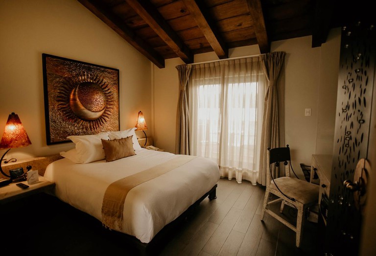 A hotel room in warm rustic tones with a large bed and copper artwork under a wood-beamed ceiling.