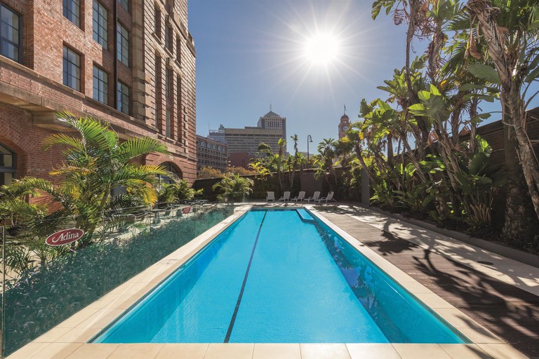 Long outdoor swimming pool framed by tropical foliage on a sunny day at Adina Apartment Hotel Sydney Central