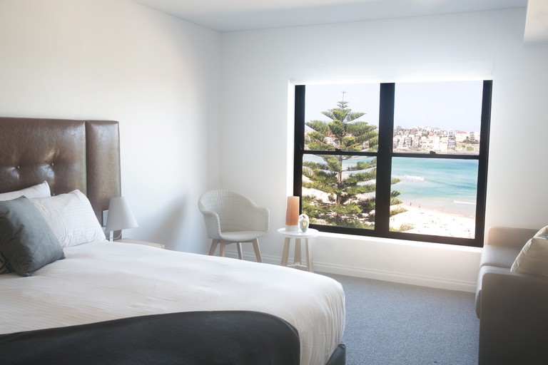 Cosy bedroom with large bed, couch, rattan chair and large picture window with beach view at Bondi 38 Serviced Apartments