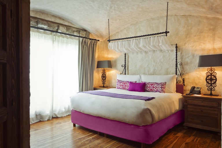 Double bed with deep pink cushions in rustic, cave-like room with ornamental lamps and a large window at Busué.