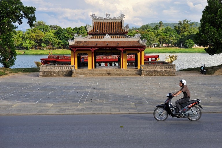 A person riding a motorbike past a pagoda next to the Perfume River in Hue