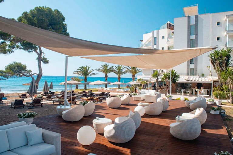 Beachfront deck with low white rattan chairs under shade sails with turquoise water beyond it at Hotel Melbeach and Spa, Mallorca