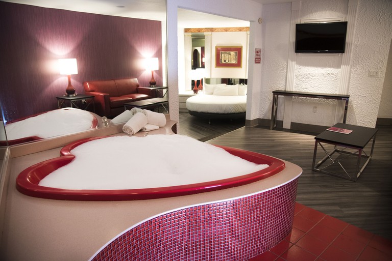 A heart-shaped hot tub with bubbles, a reflection of a couch in a mirror, and a round bed in the other area, in a hotel room at Paradise Stream Resort