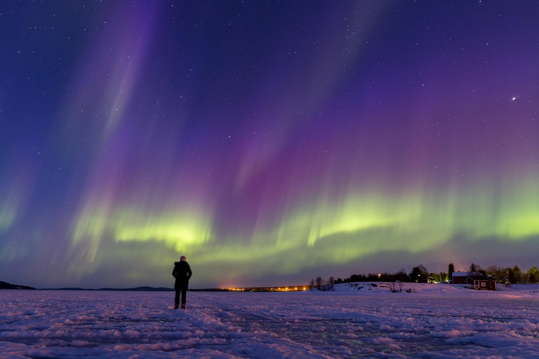 Person watching a breath-taking display of colorful northern lights on frozen lake Inari, Finland.