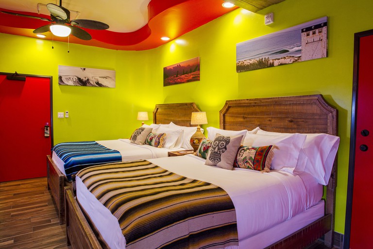 Two twin beds with Mexican-themed decorative pillows in a a colorful private room with lime-green walls, a red door and red ceiling details at Nomads Hotel