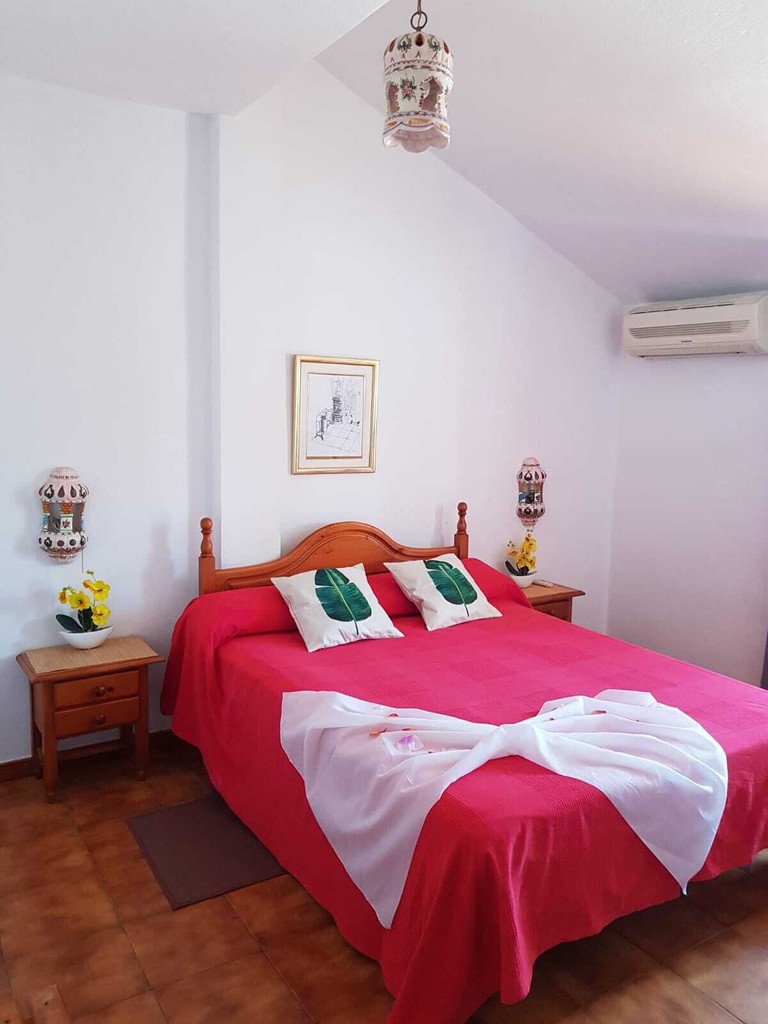 A bedroom at El Tio Mateo, with a pink bed and two leaf-print pillows, bedside tables with yellow plants and a framed picture