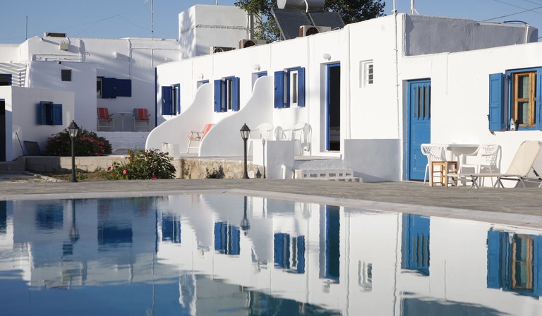 The whitewashed exterior and blue shutters of Nikos Rooms reflected in the outdoor pool