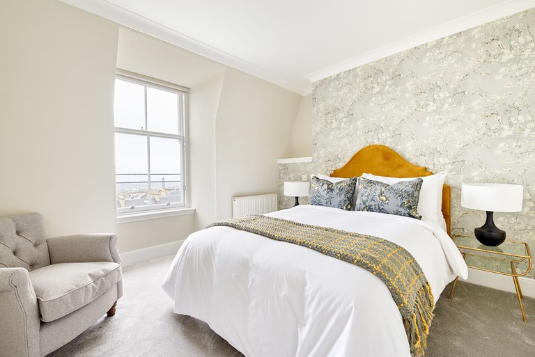 A light bedroom at Sonder – Royal Garden Apartments with a cushioned bed, an armchair, bedside tables, lamps and a window