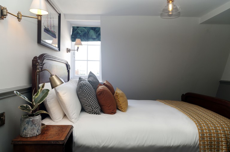 Room at Admiral Hardy has multiple pillows in different colours and patterns, a snake plant in a marble pot, a wooden bedside table and grey walls