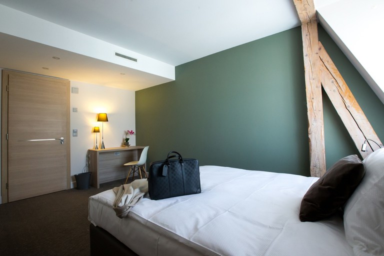 A cosy bedroom at Hôtel des Voyageurs, with a large bed, moss-green accent wall, rustic exposed beam, small desk and dark carpet