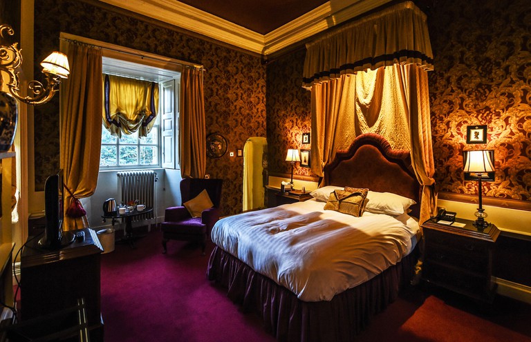 Double bed in low-lit, opulent room with armchair, TV, radiator and purple carpet at Lumley Castle Hotel