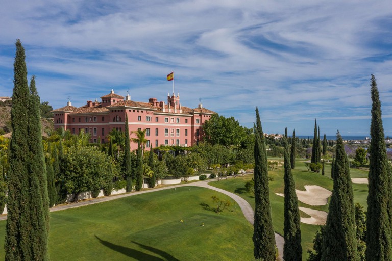The pink-hued Anantara Villa Padierna Palace Benahavís Marbella Resort surrounded by a golf course and trees on a sunny day