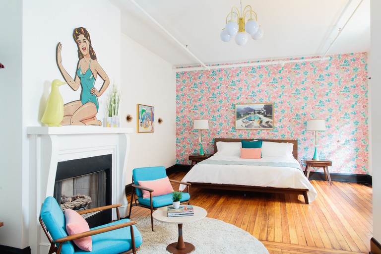 An arty bedroom with a double bed pushed up against a pastel, floral wallpaper, with hardwood floors, two turquoise armchairs with pink pillows in front of a fireplace and an artwork featuring the silhouette of a woman above the fireplace at Dwell Hotel